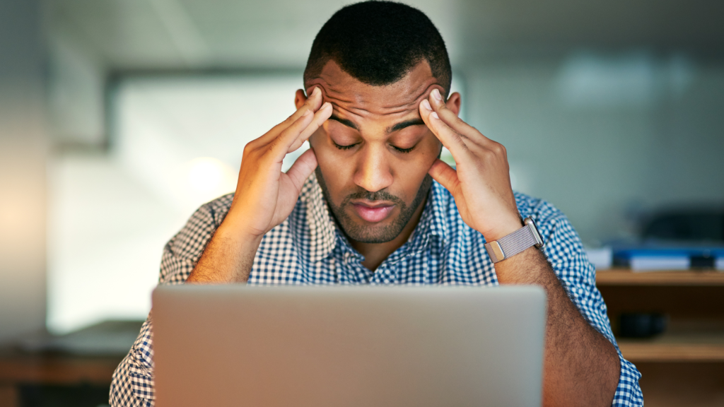 Managing conflict in the workplace. A man holding his temples looking stressed. He has a blue and white checked shirt, short haircut and is wearing a watch. Looking at a screen.